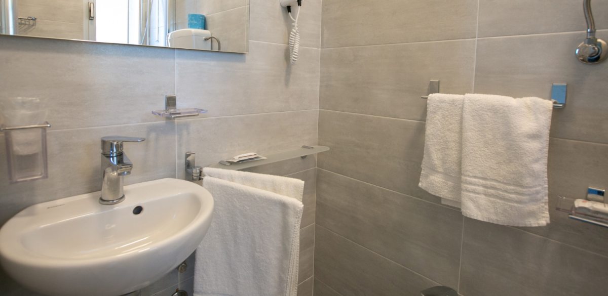 Bagno moderno con lavabo, specchio, asciugacapelli e asciugamani bianchi su piastrelle grigie.