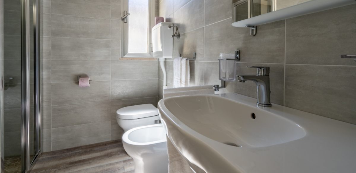 Bagno moderno con piastrelle grigie, lavabo, bidet, doccia e finestra.