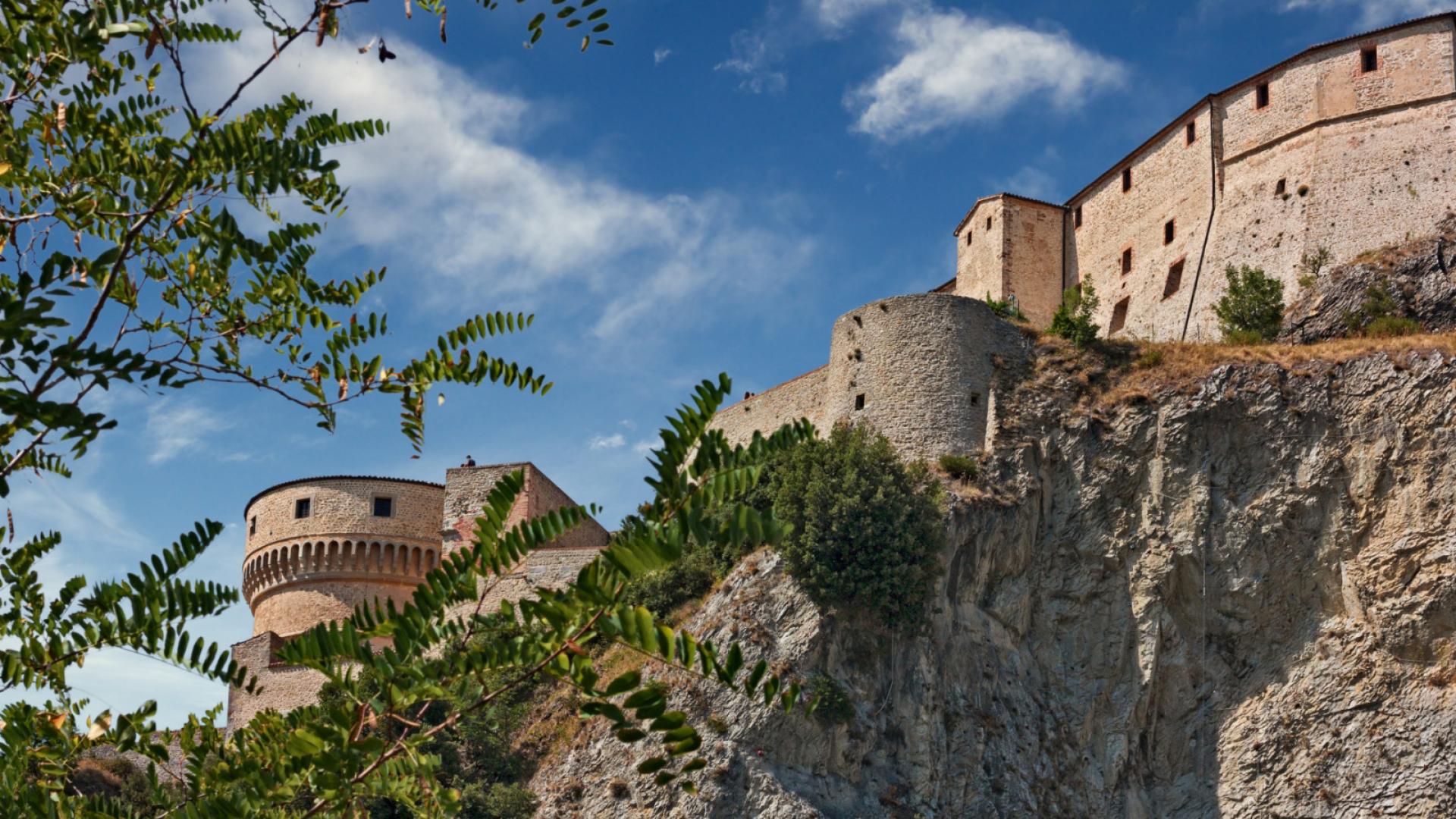 Imponente fortezza medievale su una scogliera, circondata da vegetazione e cielo azzurro.