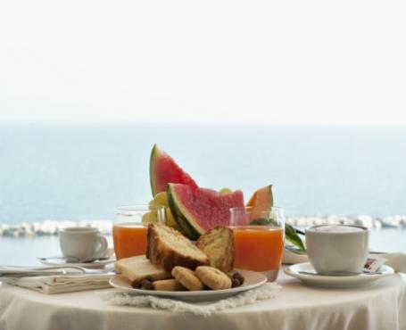 Colazione con vista mare, frutta fresca, succhi e dolci su un tavolo apparecchiato.