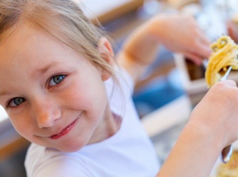 Bambina sorridente mangia spaghetti con entusiasmo, indossando una maglietta bianca.