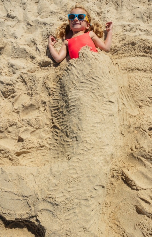 Bambina sorridente sepolta nella sabbia con coda di sirena, indossa occhiali da sole blu.