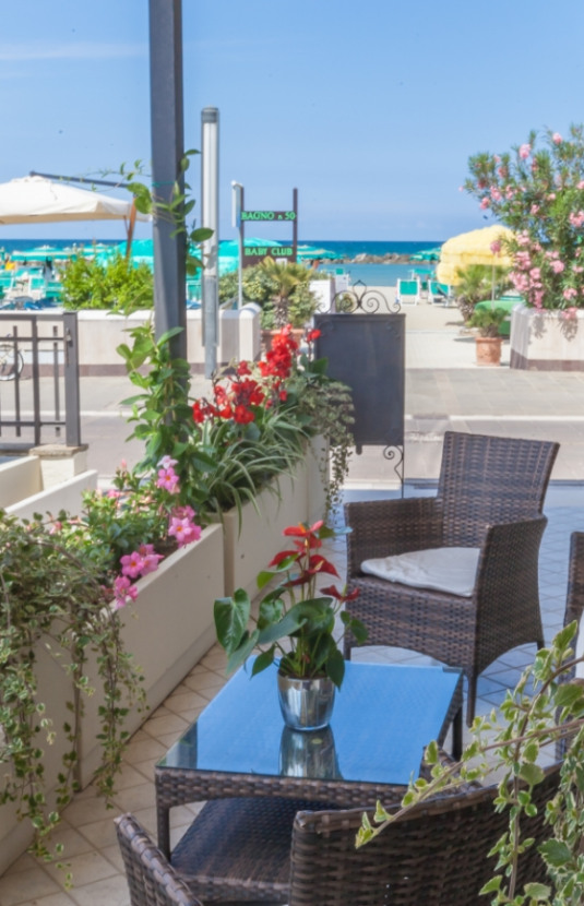 Terrazza con sedie in vimini, fiori colorati e vista mare.