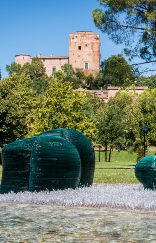 Scultura in vetro verde con fontana, parco e torre storica sullo sfondo.